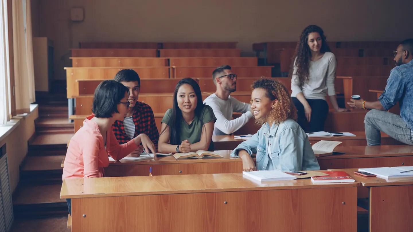 Estudiantes adultos conversando entre las bancadas de un aula universitaria con luz natural